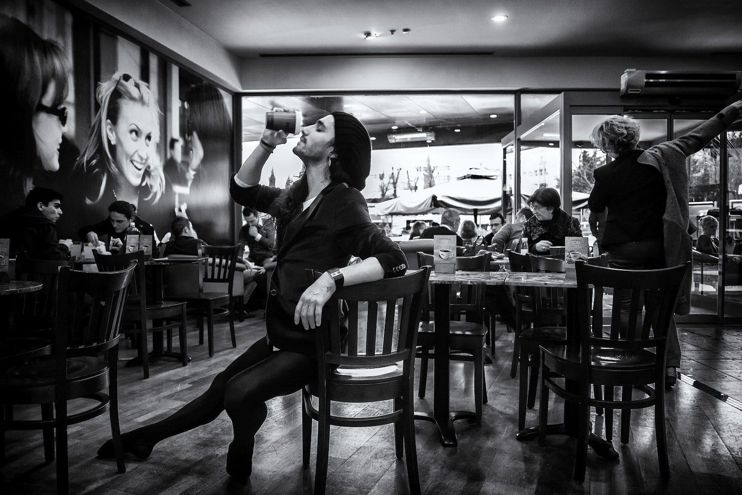 Nude Art Photography in Urban Cafés – Burak Bulut Yıldırım A figure in a café evokes contemplation amidst urban life. A person in a black outfit sips from a cup, seated in a café surrounded by patrons, with large black-and-white portraits on the wall.