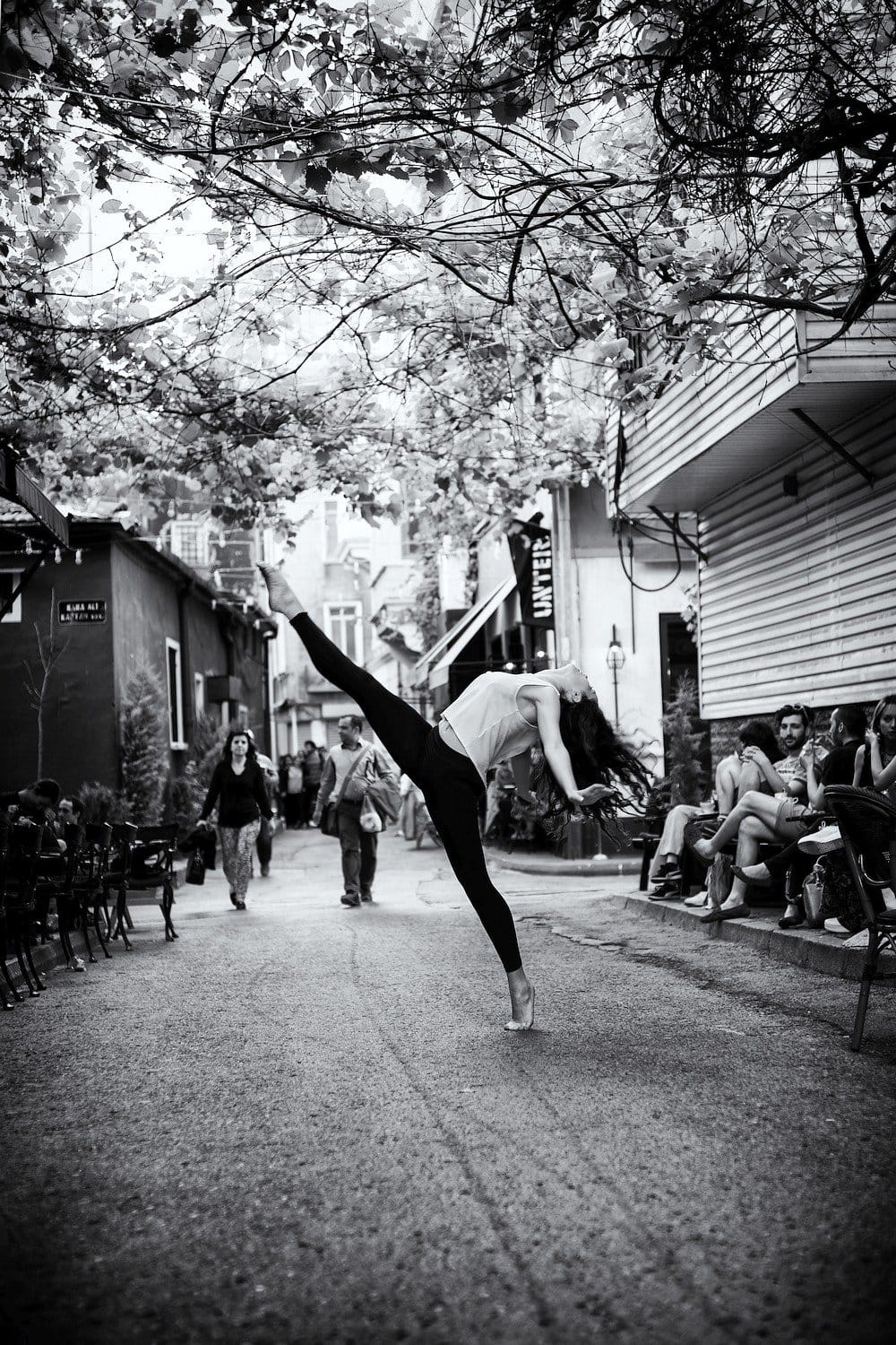 “Dancer in Motion Amidst Urban Life – Black and White Photography” A dancer in mid-pose stretches in a narrow street, surrounded by greenery, with onlookers and café seating in black and white.